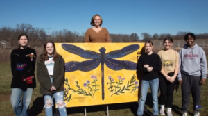 Photo of teacher with students in front of a large painting of a dragonfly.